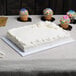 A white cake on a table with cupcakes and a donut on top, on a white Enjay 1/4 sheet cake board.