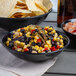 A black Vollrath melamine bowl filled with black bean and corn salsa on a table with chips and a bottle of beer.