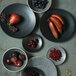 Oneida Urban by 1880 Hospitality black porcelain coupe plates on a table with bowls of fruit.