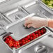 A gloved hand using a Choice stainless steel slotted steam table pan cover to pick up tomatoes from a tray.