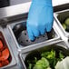 A person in blue gloves using a Vollrath stainless steel steam table pan false bottom to remove food from a metal container.