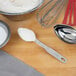 A stainless steel measuring spoon with a white powder on it next to a bowl of flour and a whisk.