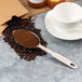 A Thunder Group stainless steel measuring scoop filled with ground coffee next to a cup and saucer.