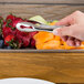A hand using American Metalcraft stainless steel tongs to serve fruit from a tray.