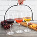 A hand holding a Vollrath glass rack with three wine glasses filled with red, yellow, and red liquid next to grapes.