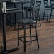 A black metal Lancaster Table & Seating cross back bar stool with a black vinyl seat next to a table with chairs.