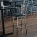 A Lancaster Table & Seating clear coat finish cross back bar stool with a black vinyl padded seat next to a table with more chairs.