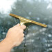 A hand holding a Unger brass window cleaner handle with a gold metal clip.