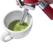 A hand using an iSi stainless steel decorating tip to pour green tea into a white cup.