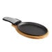 A black oval cast iron fajita skillet with a wooden underliner and a grey handle on a wooden board.