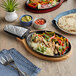A Choice oval cast iron fajita skillet with a wood underliner and a grey silicone handle cover on a table with sizzling fajitas.