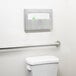 A Lavex stainless steel surface mounted toilet seat cover dispenser on a wall above a toilet with a green sign.