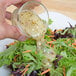 A hand pouring Ken's Foods Greek Vinaigrette with Feta Cheese and Black Olives dressing onto a bowl of salad.