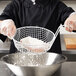 A person in a chef's uniform using a Thunder Group coated wire basket to pour flour into a bowl.