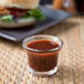 A small glass cup of Ken's Cannonball BBQ Sauce on a table next to a burger.