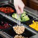 A person holding a Cambro clear plastic perforated salad bar spoon over a tray of food.