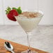 A close up of a martini glass of white tapioca pudding with a strawberry on top with a spoon on a wood surface.
