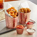 A copper Acopa French fry holder filled with fried food on a table with a bowl of red sauce.
