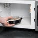 A hand holding a Pactiv black oval microwavable container of food in a microwave.