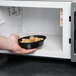 A hand holding a Pactiv Newspring VERSAtainer oval black bowl of food in a microwave.