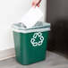 A hand putting a piece of paper into a green Rubbermaid recycling bin.