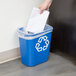 A hand putting a piece of paper into a blue Rubbermaid recycling bin with a white recycle symbol.