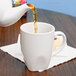 A white teapot pouring tea into a Tuxton white bistro mug.