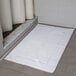 An Oxford Reserve white bath mat with a dobby border on a tile floor.