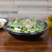 A bowl of salad with a clear plastic dome lid on a table.