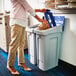 A woman putting a bag into a white rectangular Rubbermaid recycling bin with open and mixed recycling lids.