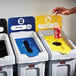 A hand putting paper into a blue Rubbermaid recycling container with a black rectangular lid.