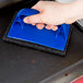A hand using a blue Scrubble grill polish pad to clean a black surface.