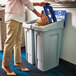 A woman putting a box into a blue Rubbermaid Slim Jim recycling bin with a white rectangular lid.