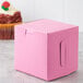 A 4" x 4" pink bakery box with a cupcake inside and a handle.