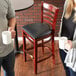 A man and woman holding coffee cups in front of a Lancaster Table & Seating mahogany wood bar stool with black vinyl seat.