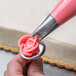A person using an Ateco rose piping tip to pipe pink frosting flowers on a cake.
