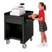 A girl in a red sweater and plaid skirt standing next to a black Cambro cash register stand with trays on both sides.