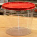 A clear plastic Carlisle food storage container with a red lid on a table.