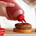 A person using a Vollrath Color-Mate Squeeze Bottle with a red cap to pour ketchup onto a burger.
