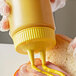 A person using a Vollrath Twin Tip Squeeze Bottle with a yellow cap to put mustard on a sandwich.