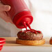 A hand using a Vollrath Clear Twin Tip Squeeze Bottle with a red cap to pour ketchup on a burger.
