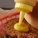 A person pours yellow mustard from a Vollrath Traex bottle onto a burger.
