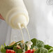 A person using a Vollrath clear wide mouth squeeze bottle to pour dressing on a salad.
