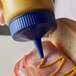 A person using a Vollrath blue wide mouth bottle cap to pour mustard on a sandwich.