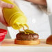 A hand using a Vollrath Tri Tip squeeze bottle with a yellow cap to pour mustard on a burger.
