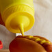 A person using a Vollrath yellow plastic bottle cap to pour mustard on a hot dog.