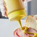 A person using a Vollrath yellow wide mouth squeeze bottle to pour mustard on a sandwich.