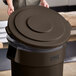 A person putting a brown Lavex commercial trash can lid on a brown trash can.