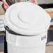A person holding a white plastic lid over a Lavex white round commercial trash can.