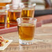 An Acopa shot glass filled with brown liquid on a table with other glasses of alcohol and a plate of nuts.